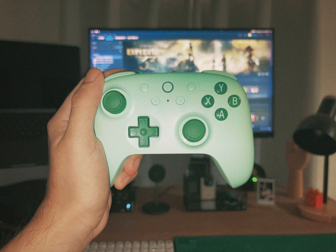 Hand holding a mint green Xbox controller in front of a gaming setup
